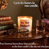 A lit candle in a clear glass jar sits on a polished wooden table, surrounded by stacked books, tortoiseshell eyeglasses, and a glass of amber liquid. White text at the top reads 'Up to 80 hours in one candle.