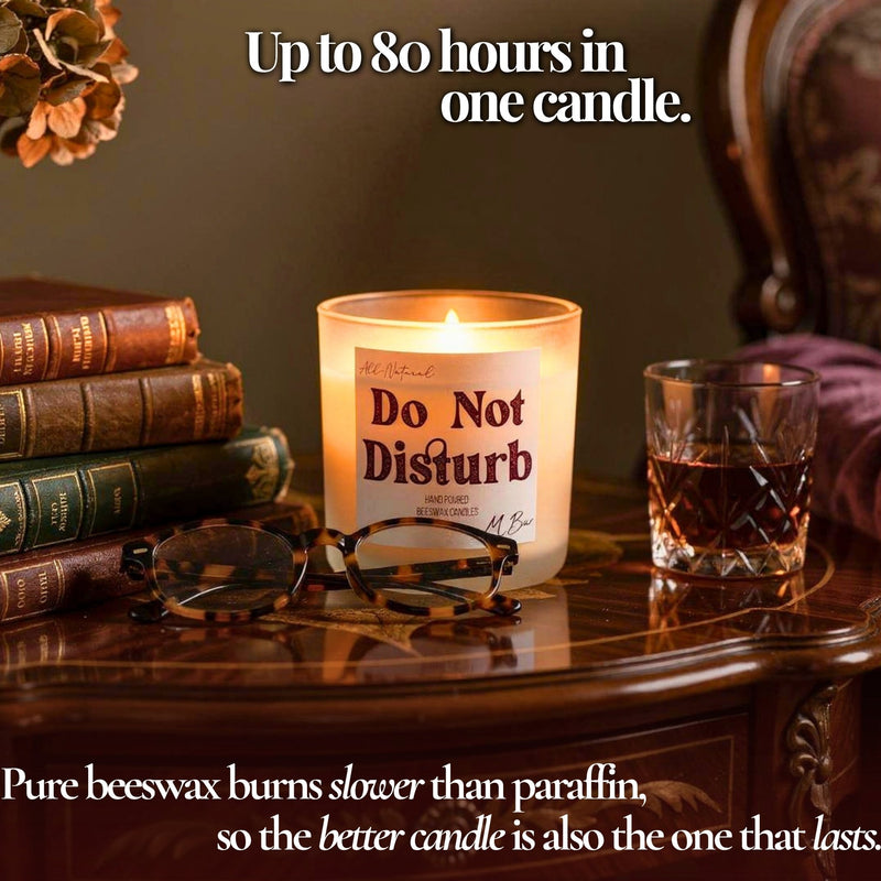 A lit candle in a clear glass jar sits on a polished wooden table, surrounded by stacked books, tortoiseshell eyeglasses, and a glass of amber liquid. White text at the top reads 'Up to 80 hours in one candle.